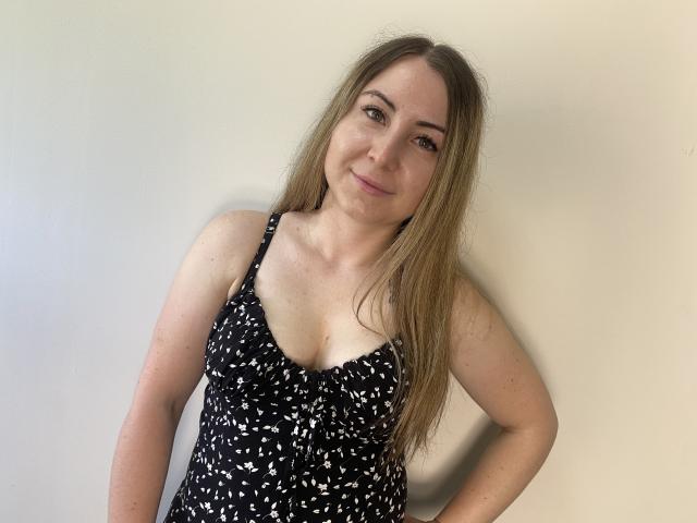 Une femme aux longs cheveux blonds, souriante, portant une robe noire et blanche à motifs avec un décolleté plongeant, qui révèle ses petites seins, debout devant un mur uni..