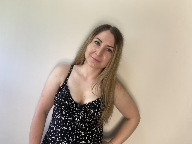 Une femme aux longs cheveux blonds, avec de petites seins, portant une robe noire avec des motifs blancs, souriant légèrement, debout devant un mur uni. Ses mains reposent sur ses hanches..