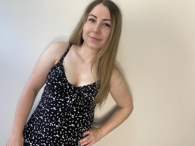 Aminas with long blonde hair, small tits, wearing a black, white-patterned dress, standing against a plain wall, smiling slightly, hand on hip, light skin, casual, cute.