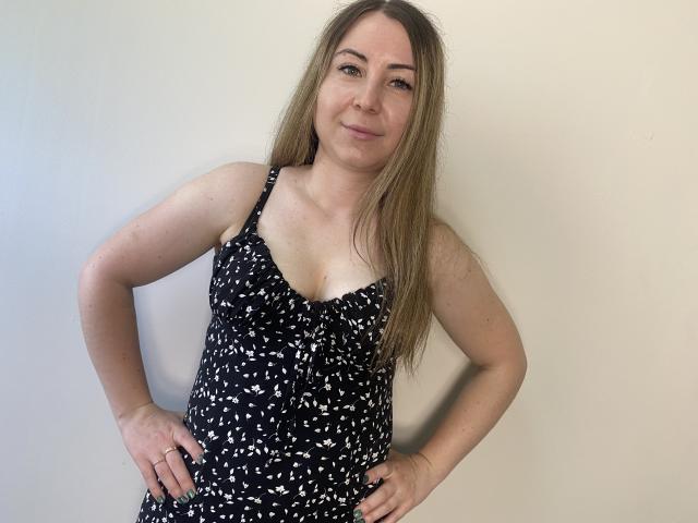 Aminas with long brown hair, small tits, wearing a black and white patterned dress, stands with hands on hips, smiling slightly, in a simple indoor setting.
