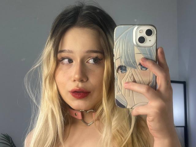 NolaMannon with blonde hair, red lipstick, and a pink heart choker, holding a phone with anime girl art, taking a selfie, showing off her tits and cute face.