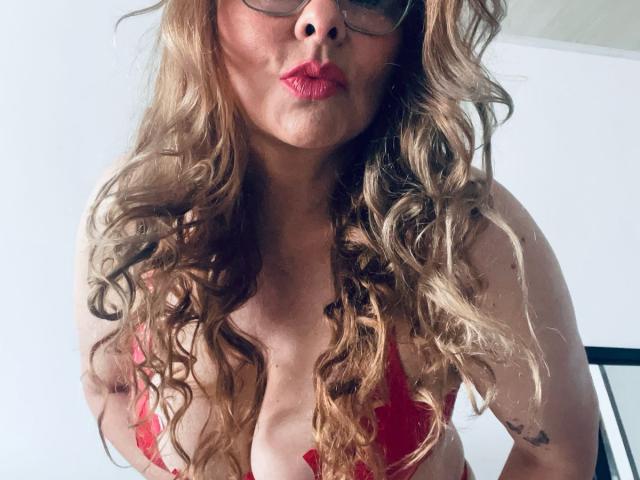 MiaAmoretti, con cabello rubio rizado, lápiz labial rojo, gafas y un sujetador de encaje rojo, mostrando sus grandes senos y un tatuaje en su brazo derecho, en un selfie..