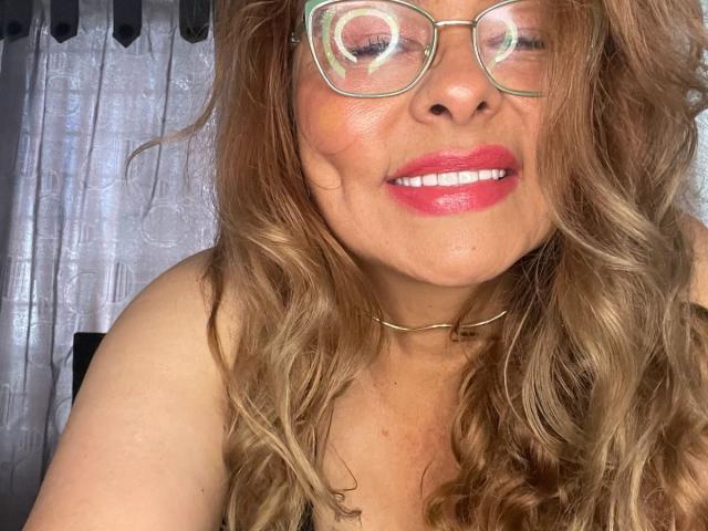 MiaAmoretti heeft golvend bruin haar, een bril, roze lippenstift en een gouden ketting, en ze glimlacht met haar ogen gesloten. Haar borsten zijn licht zichtbaar, en ze draagt een zwarte top..