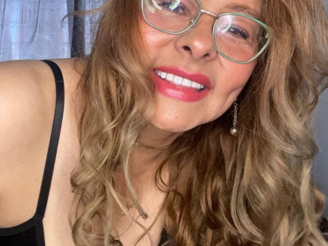 MiaAmoretti met golvend blond haar, groene bril, rode lipstick, zwarte jurk met bandjes, lachend, met tanden, parel oorbellen, zacht licht, casual selfie, bovenlichaam, schattig, aantrekkelijk..