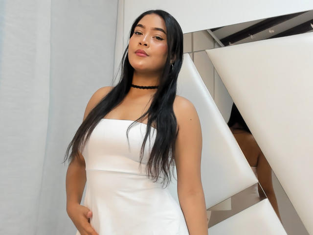 VickyRuiz, com cabelos longos e pretos, vestido branco sem alças, colar preto e maquiagem discreta, está em pé com a mão na cintura, exibindo suas nádegas, em um quarto moderno com espelhos..