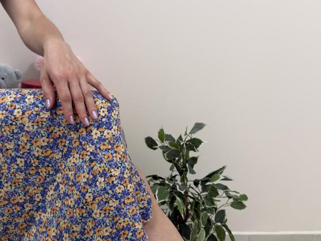 La mano di Rorria è appoggiata sulla sua gonna floreale, con fiori blu e gialli, mostrando le sue unghie dipinte di rosa. La gonna è sollevata, rivelando il suo sedere scoperto. In secondo piano, una pianta in vaso..