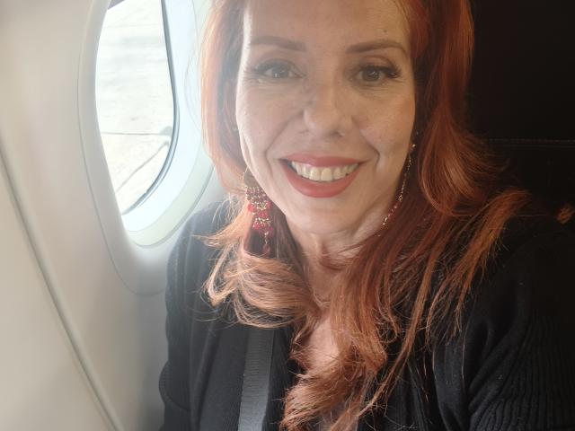 Khharha has red hair, a black shirt, and is smiling with red lipstick, wearing red earrings, seated in a car with a window to her left.