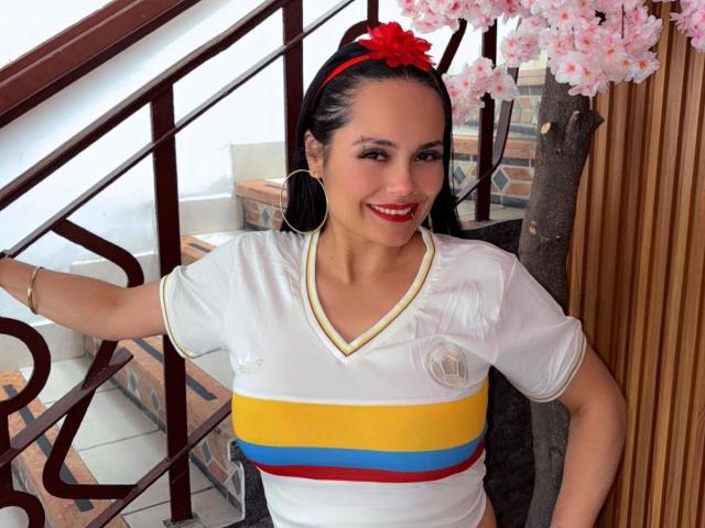 Charlotte Crofort, con cabello oscuro, una diadema de flores rojas, una camisa blanca con rayas amarillas, azules y rojas, sonriendo, aretes de aro, lápiz labial rojo, de pie junto a una escalera con flores de cerezo rosadas..