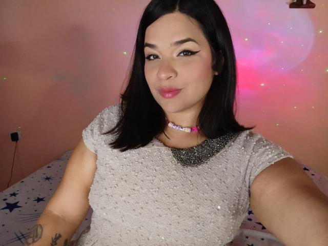 Antonela, con i capelli neri, il rossetto rosa e una collana scintillante, indossa una camicia glitterata, sta scattando un selfie, mostrando un tatuaggio sul braccio destro, con un leggero sorriso..