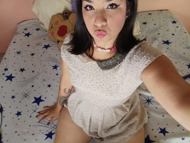 Antonela, com cabelo escuro, vestindo um vestido cinza transparente, lábios curvados, colar rosa, tatuagem no braço direito, sentada em uma cama com padrão de estrelas, com a vulva levemente visível..