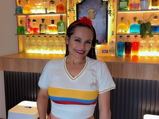 Charlotte Crofort, avec ses cheveux foncés, un bandeau rouge, un crop top blanc avec des rayures jaunes, bleues et rouges, souriante, debout devant un bar illuminé avec des bouteilles de spiritueux colorées..