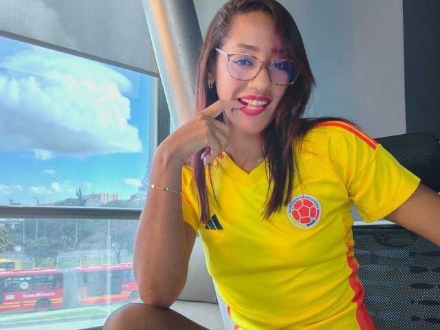 Samantha Corine mit langen braunen Haaren, Brille und rotem Lippenstift, trägt ein gelbes Fußballtrikot, zeigt auf ihren Mund und sitzt an einem Fenster, vor dem ein Bus steht..