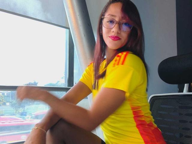 SamanthaCorine mit Brille, gelbes und rotes Hemd, sitzend, Beine gekreuzt, Blick zur Kamera, rosiger Lippenstift, langes braunes Haar, trägt ein Armband am Knöchel, sitzt auf einem Bürostuhl, im Hintergrund ein Fenster..