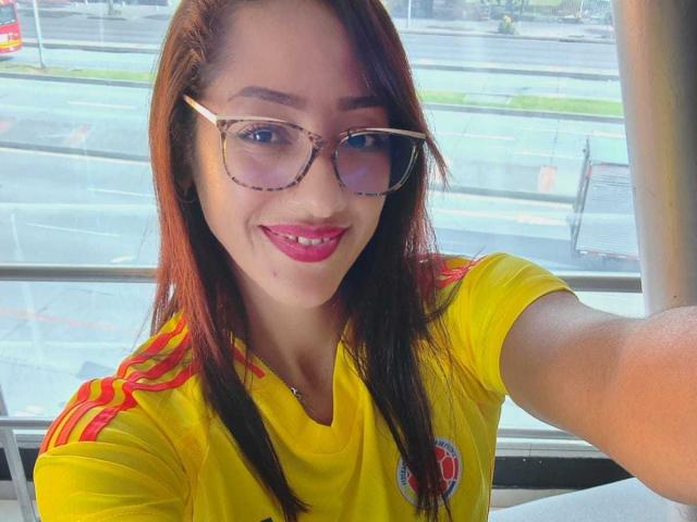 SamanthaCorine, con cabello castaño largo, vistiendo una camiseta de fútbol amarilla, gafas de montura de carey y lápiz labial rojo, sonriendo a la cámara, con los senos no visibles..