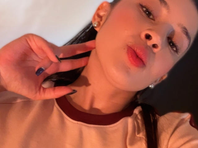EmmaLombardi with pouty red lips, dark nails, and a hand near her neck, wearing a beige shirt with red trim. She has a light skin tone and brown hair.