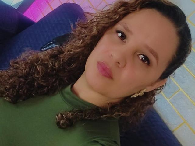 VittoriaI tem cabelos castanhos cacheados, olhos castanhos e batom rosa. Ela está vestindo uma blusa verde e brincos dourados, olhando para a câmera com uma expressão levemente emburrada..