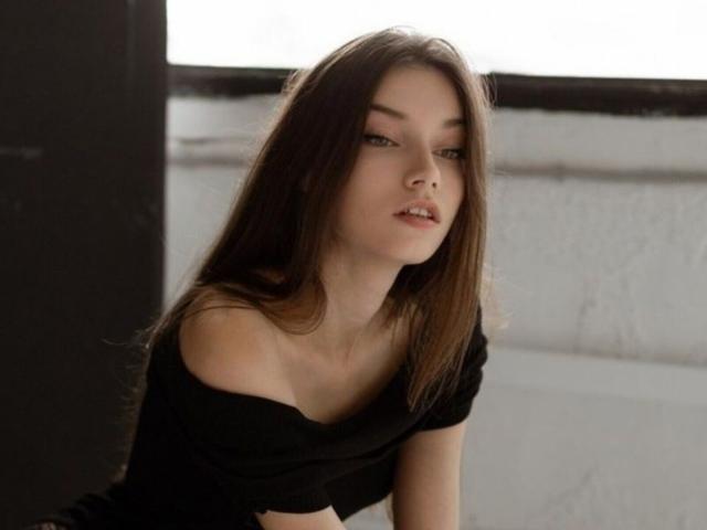Leanne Yellowhair, avec ses longs cheveux bruns, sa peau pâle et ses yeux verts, porte un haut noir à une épaule, et regarde légèrement sur le côté avec une expression subtile et légèrement boudeuse..