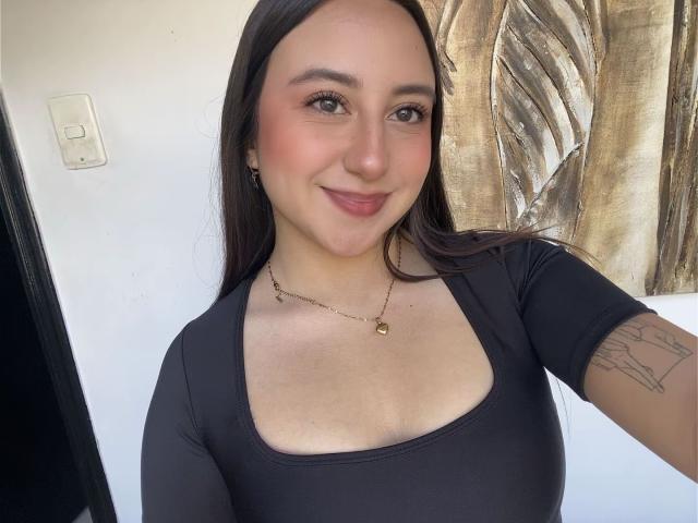 Rosalie Dupont, com cabelos castanhos longos, blusa preta, colar de ouro pequeno, tatuagem no braço direito, sorrindo, pele clara, olhos castanhos, bochechas rosadas, seios de tamanho médio, parede branca, obra de arte dourada..