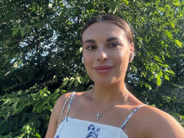 BlackFatal with brown hair, green eyes, and a small cross necklace, wearing a white floral dress, smiling with a subtle sunlit glow on her fair skin, outdoors.