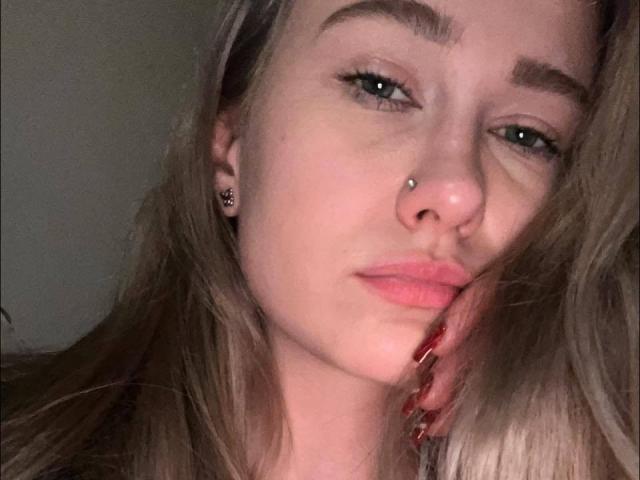 GlossyBambe, com cabelo loiro claro, olhos azuis e um piercing no nariz, olhando ligeiramente para o lado, com unhas pintadas de vermelho tocando sua bochecha. Ela tem uma expressão sutil e levemente emburrada..