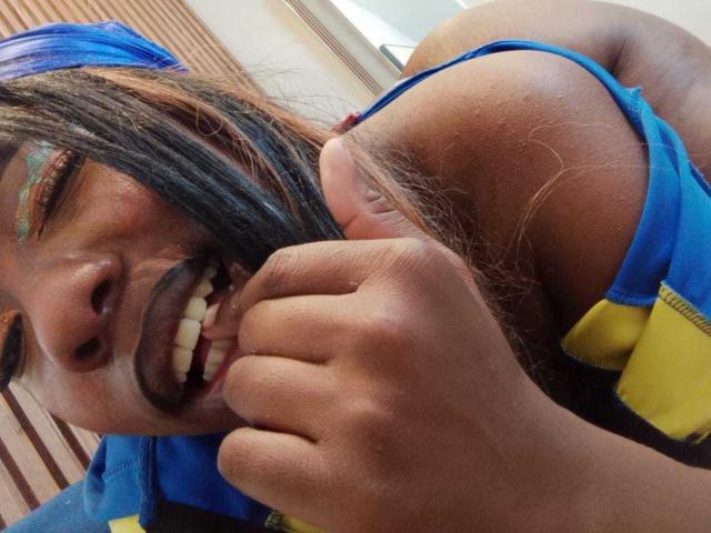 JazaraKoral mit dunkelbrauner Haut, einem blauen Tanktop und einem gelben Reißverschluss, mit schwarzen Haaren, lächelnd mit sichtbaren Zähnen, mit grünem Lidschatten und einem blauen Kopftuch, hält ihre Haare in der Hand..