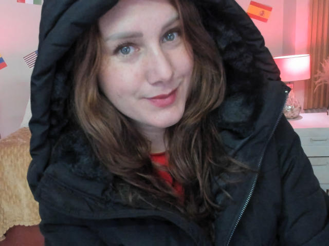 KatyaRogersX mit blauen Augen, welligem braunen Haar und einer schwarzen, gepolsterten Jacke, lächelt in die Kamera und zeigt einen Hauch eines roten Hemdes darunter..
