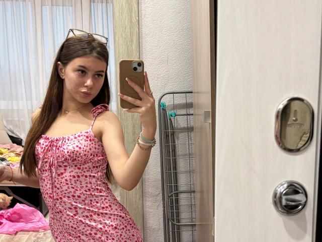 KimberlyZampieri with long brown hair, wearing a pink and white heart-patterned dress, holding a phone, showing her mirror selfie, with a serious expression, and a small ass.