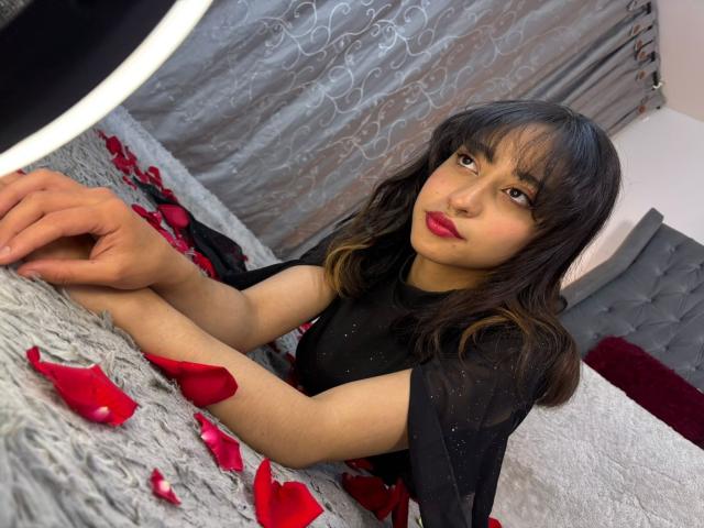 CamiVargas, met een medium bruine huid, zwart golvend haar, rode lippenstift en een zwarte jurk, ligt op bed met rozenblaadjes, kijkt naar de camera en heeft haar handen op een grijze, gestructureerde deken..