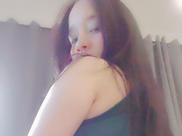 ValeriaRossii with long brown hair, pale skin, and subtle makeup, looking over her shoulder, showing her bare shoulder and black strap top, in a softly lit room.