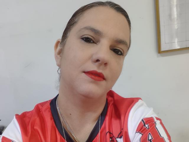 Amelia Wilson tem pele clara, batom vermelho, delineador preto e está vestindo uma camisa esportiva vermelha e branca com um colar de corrente. Seu cabelo castanho está preso para trás..