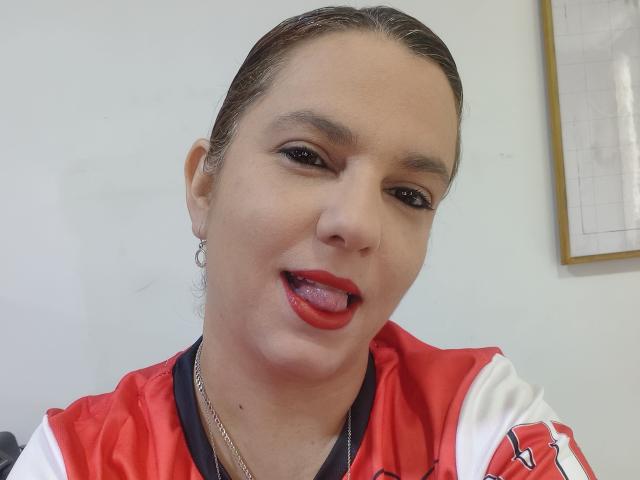 Amelia Wilson, com cabelo castanho, usando uma jaqueta esportiva vermelha e branca, batom vermelho, língua de fora, brincos pequenos e redondos e um colar de prata, olhando para a câmera..