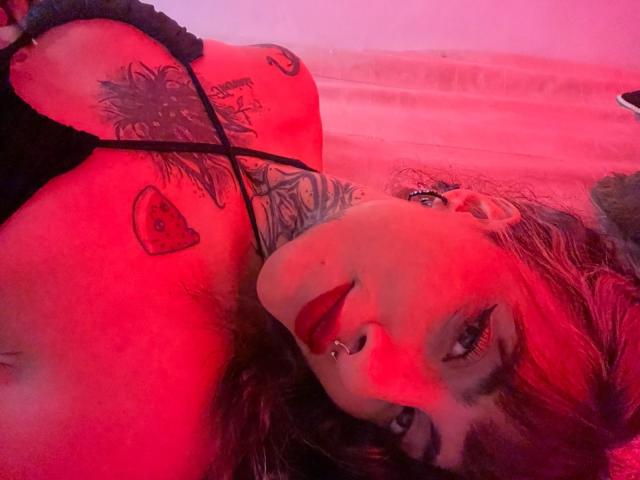 LiaMiss with red-tinted skin, black lace top, tattoos on chest and neck, nose ring, and red lipstick, lying down, looking at camera, red lighting, dark wavy hair.