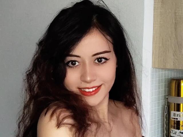 Jettie Korczynski, met lang, golvend zwart haar, rode lippenstift, en met een glimlach, toont haar blote schouders en lieve gezicht, terwijl ze naast een houten kast met een blikje staat..