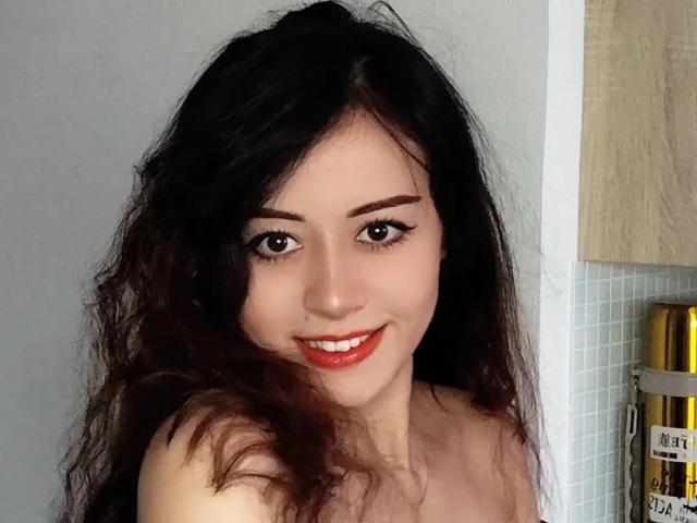 Jettie Korczynski, com cabelos longos, escuros e ondulados, sorrindo, batom vermelho, delineador preto e fundo branco. Ela está sem blusa, mostrando os ombros nus. Suas nádegas não estão visíveis..