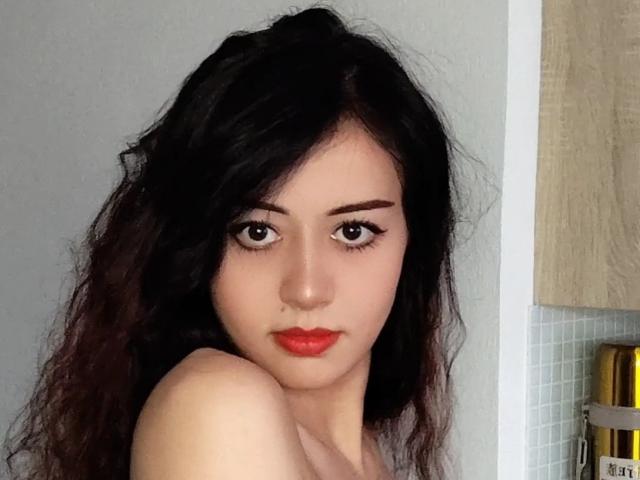 Jettie Korczynski heeft donker, golvend haar, bleke huid, rode lippenstift en dikke zwarte eyeliner. Haar blote schouders zijn zichtbaar. Ze heeft een serieuze, enigszins verleidelijke uitdrukking..
