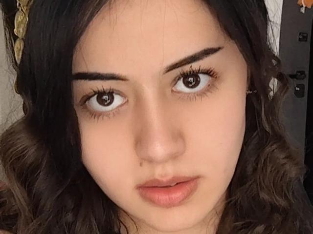 Jettie Korczynski tem cabelos escuros e ondulados, sobrancelhas grossas, olhos castanhos e lábios rosados levemente entreabertos. Ela olha diretamente para a câmera com uma expressão neutra..