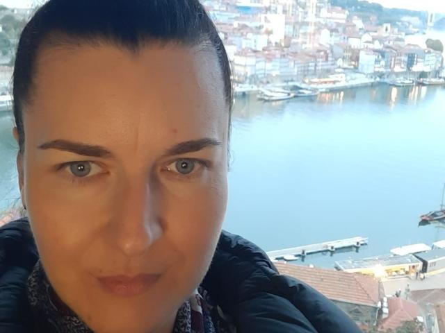NadiaRose with brown hair in a ponytail, blue eyes, and a pouty expression, wearing a black jacket, staring into the camera, with a waterfront city in the background.