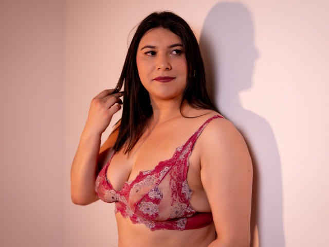 ZoeyGrey, with medium tits, wearing a red lace bra, stands against a white wall, looking to the side, dark brown hair, slight smile, right hand touching hair.