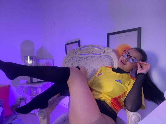IrisBlair, with glasses, sits on an ornate chair, legs up, wearing a yellow soccer shirt, black mesh sleeves, and black knee socks, exposing her bare pussy.