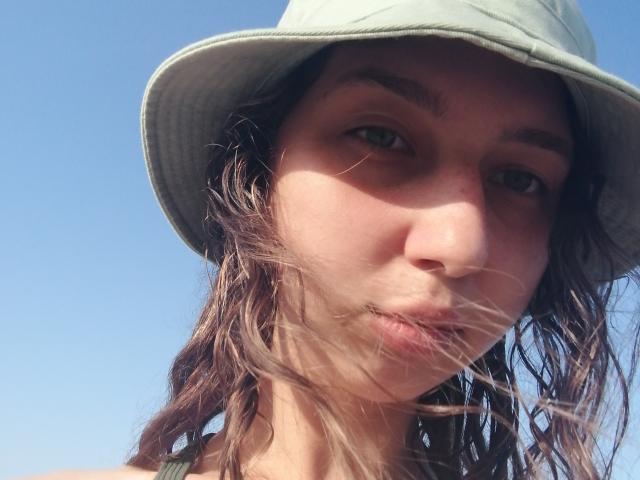 Sanella's face with a green hat, brown wavy hair blowing in the wind, and slightly pouty lips. Her light green eyes look directly at the camera.