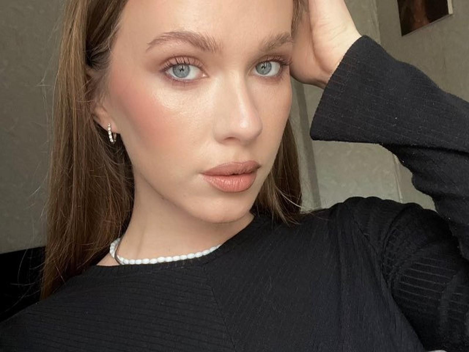 Kalyn Ivie, met blauwe ogen, draagt een zwarte geribbelde top, een witte parelketting en kleine oorbellen. Ze heeft haar hand in haar bruine haar en kijkt serieus, met lichtroze lippen..