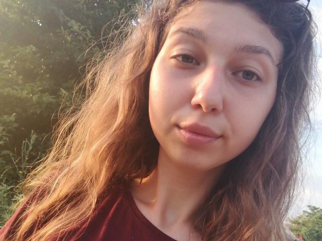 Sanella has wavy brown hair, light skin, and a small nose. She's wearing a red shirt, looking at the camera with a slight smile. Her pussy is not visible.
