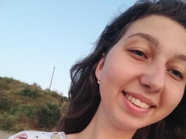 Sanella's smiling face with wavy dark brown hair, light skin, small earrings, and cute teeth. She's wearing a floral top, and her nose is slightly upturned.