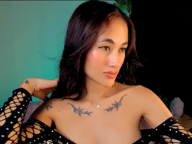 HarleyRoods with dark hair, wearing a black fishnet top, showing tattoos on her chest, and a small star tattoo on her left hand, smiling slightly. Her tits are visible.