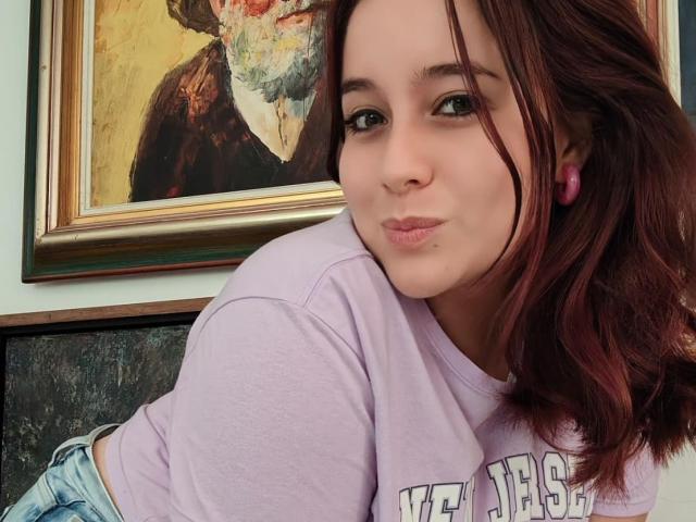 AngelaMonticelli with pouty lips, pink earbuds, light purple shirt, and brown wavy hair, leaning on a painted portrait of a person with a colorful face.