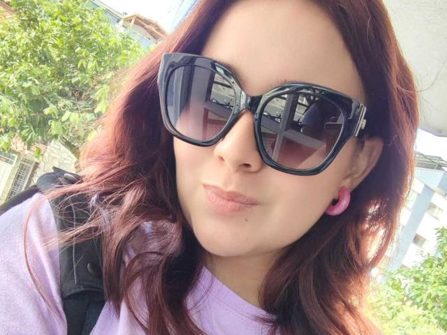 AngelaMonticelli with red hair, wearing black sunglasses and pink ear gauges, purple shirt, and black backpack, pouty lips, slight smile, sunlit, outdoor setting.