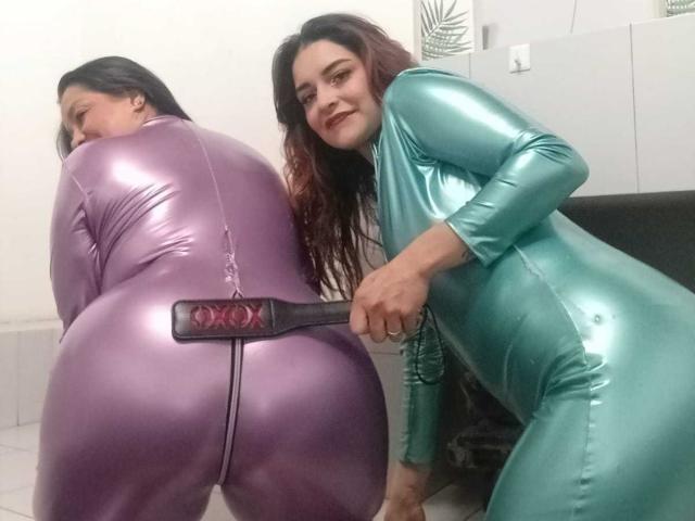 SaraAndMarilyn69 vestindo bodies de látex roxo e verde brilhantes, ajoelhadas com os traseiros levantados, um plug anal preto no ânus da que veste o roxo, sorrindo para a câmera..