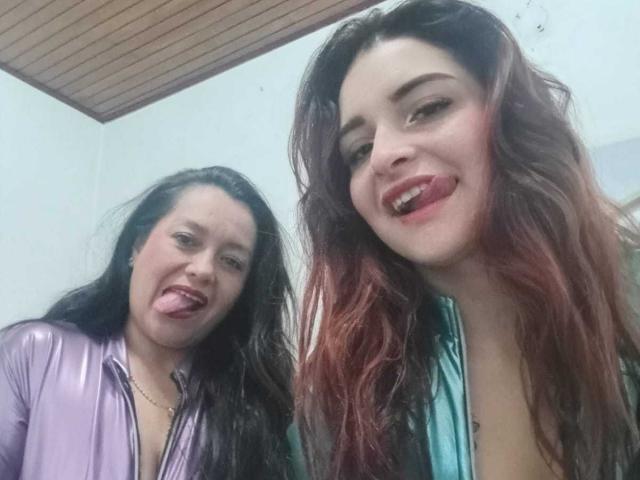SaraAndMarilyn69 avec de longs cheveux noirs, un haut violet brillant, la langue tirée et un sourire. SaraAndMarilyn69 avec des cheveux teints en rouge, un haut vert, la langue tirée et un sourire. Les deux ont de petits seins..