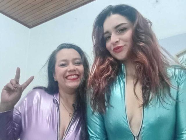 SaraAndMarilyn69 in shiny purple and green outfits, purple top showing cleavage, green top showing deep cleavage, smiling, making peace sign, red lipstick, wavy brown hair.