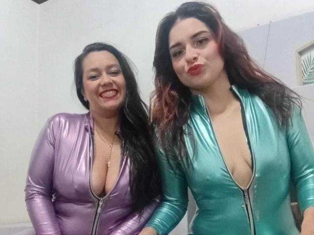 SaraAndMarilyn69 mit großen Brüsten in glänzenden, lila und grünen, vorne zu öffnenden Bodys, lächelnd und posierend. Beide haben langes, schwarzes Haar, roten Lippenstift und freiliegende Oberteile..
